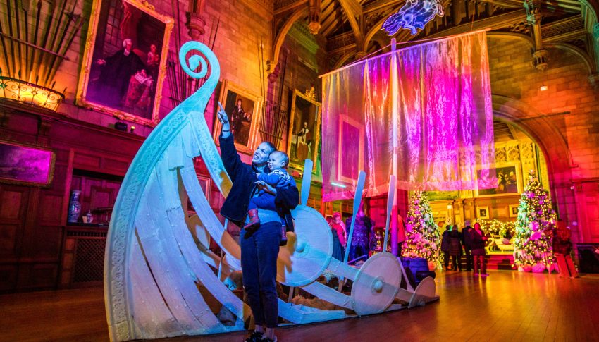 Parent holding child as they point up to the ceiling of a grand and colourful room. There is a groovy-looking pirate ship installation in the middle of the room.