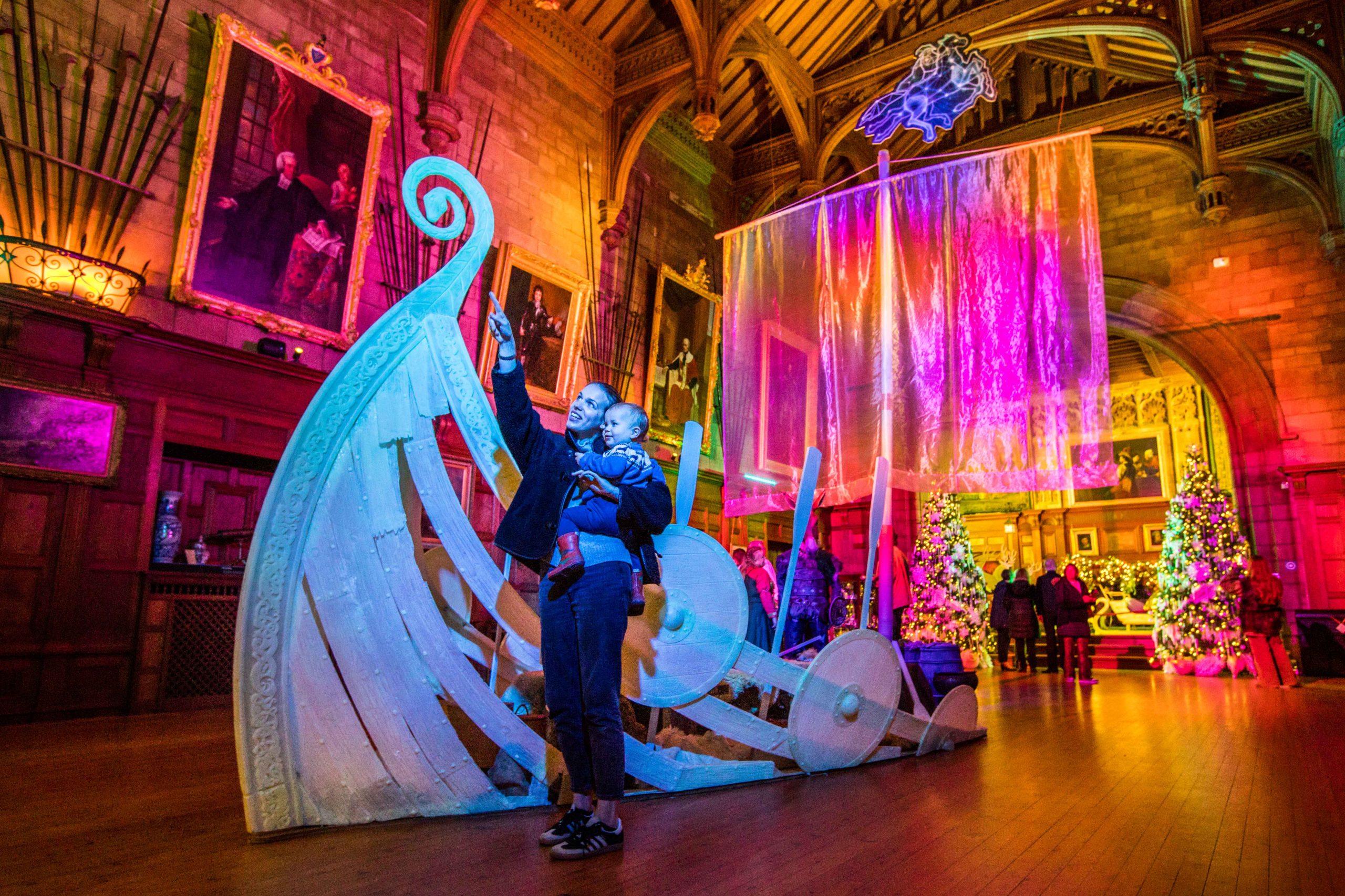 Parent holding child as they point up to the ceiling of a grand and colourful room. There is a groovy-looking pirate ship installation in the middle of the room.