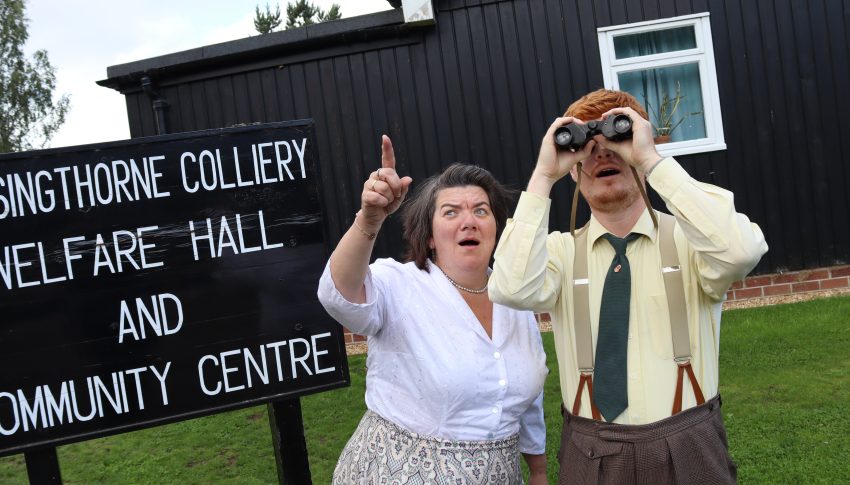 Two people in Beamish Museum costume and outside the Welfare Hall one looking up at the sky with binoculars and the other pointing in the distance.