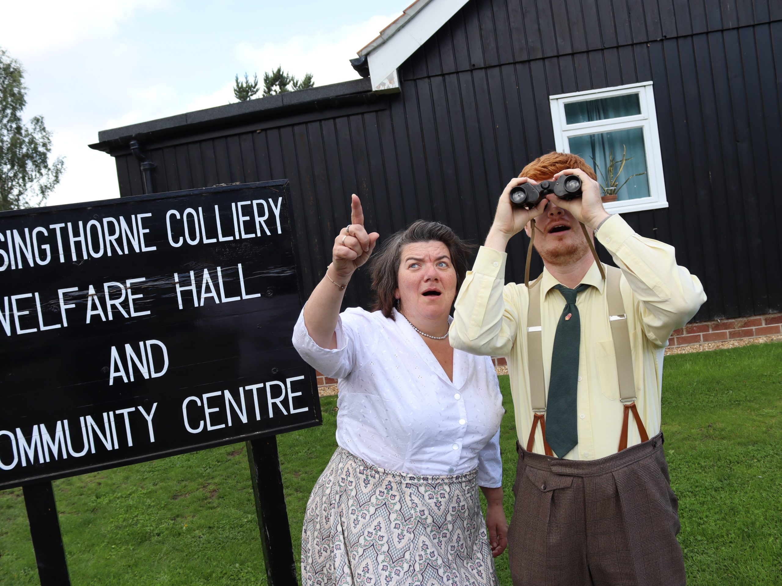 Two people in Beamish Museum costume and outside the Welfare Hall one looking up at the sky with binoculars and the other pointing in the distance.