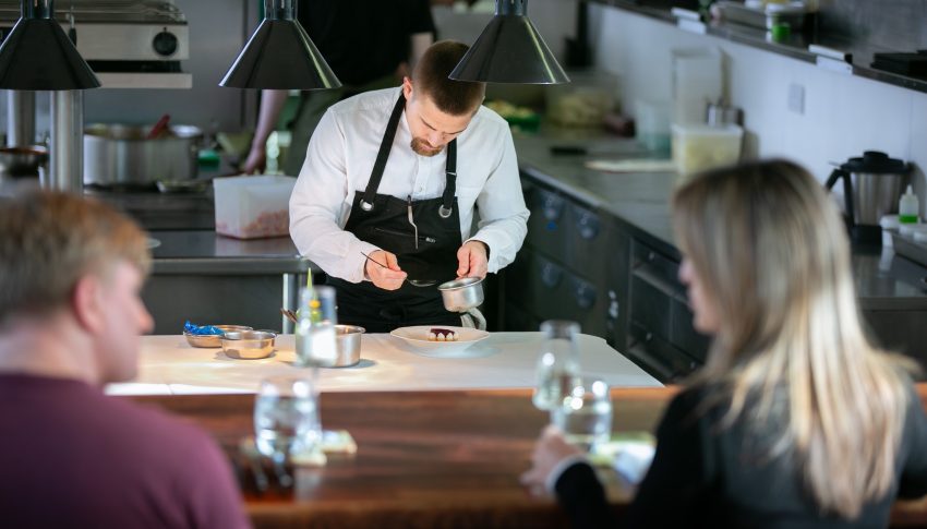 Chef adding finishing touches to a plate with customers looking on in the foreground.
