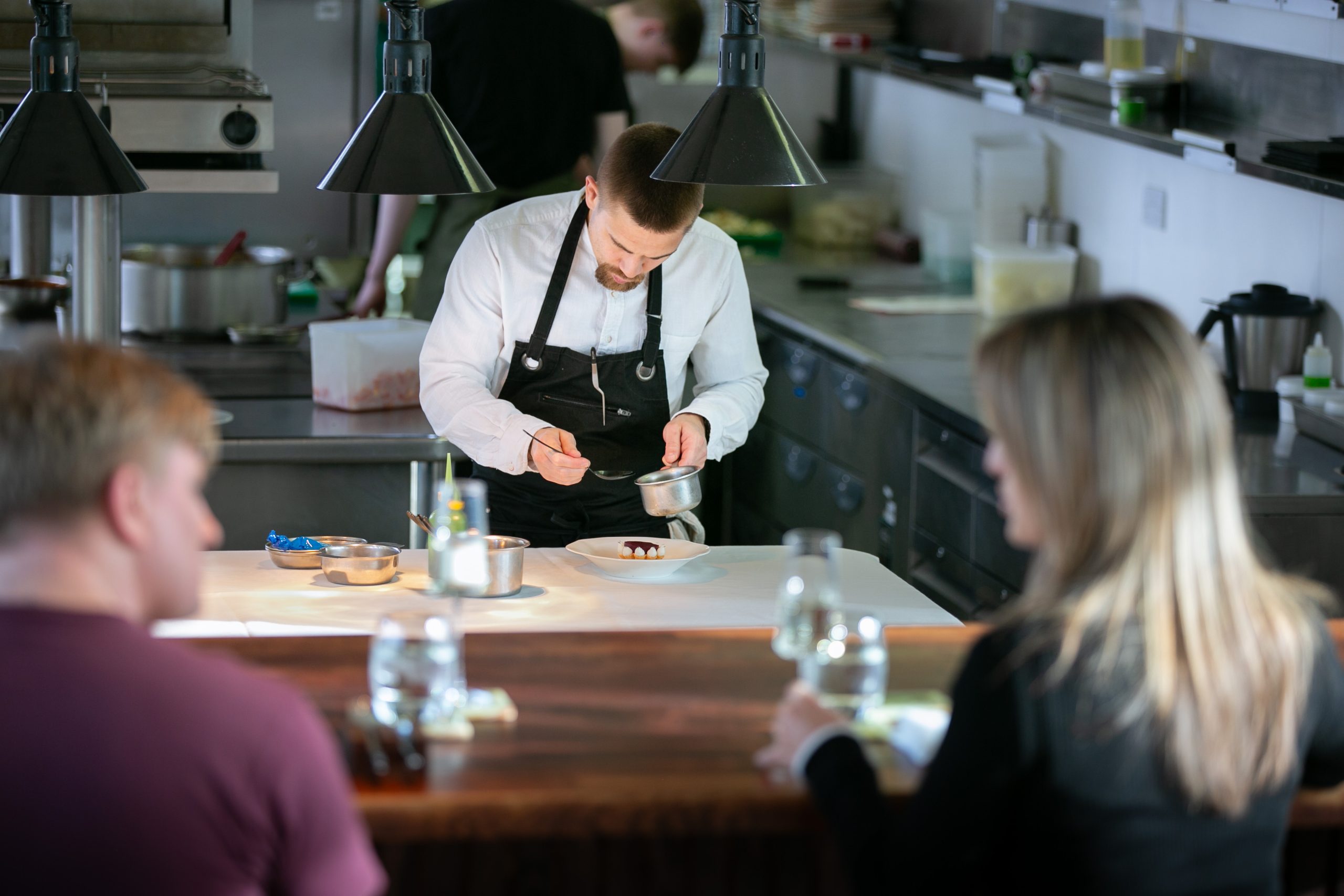 Chef adding finishing touches to a plate with customers looking on in the foreground.