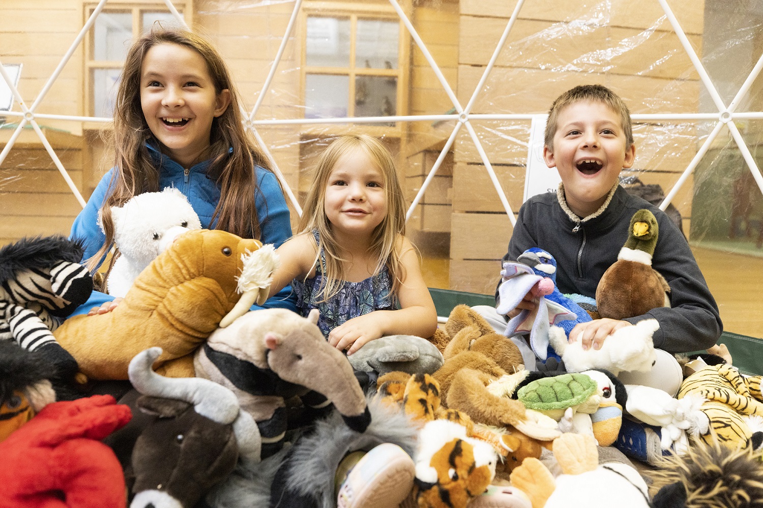 Three children sat happily with lots of stuffed animal toys smiling to camera.