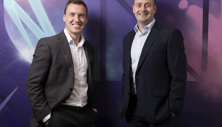 Danny and Stephen stood together against a colourful spray painted wall smiling to camera.