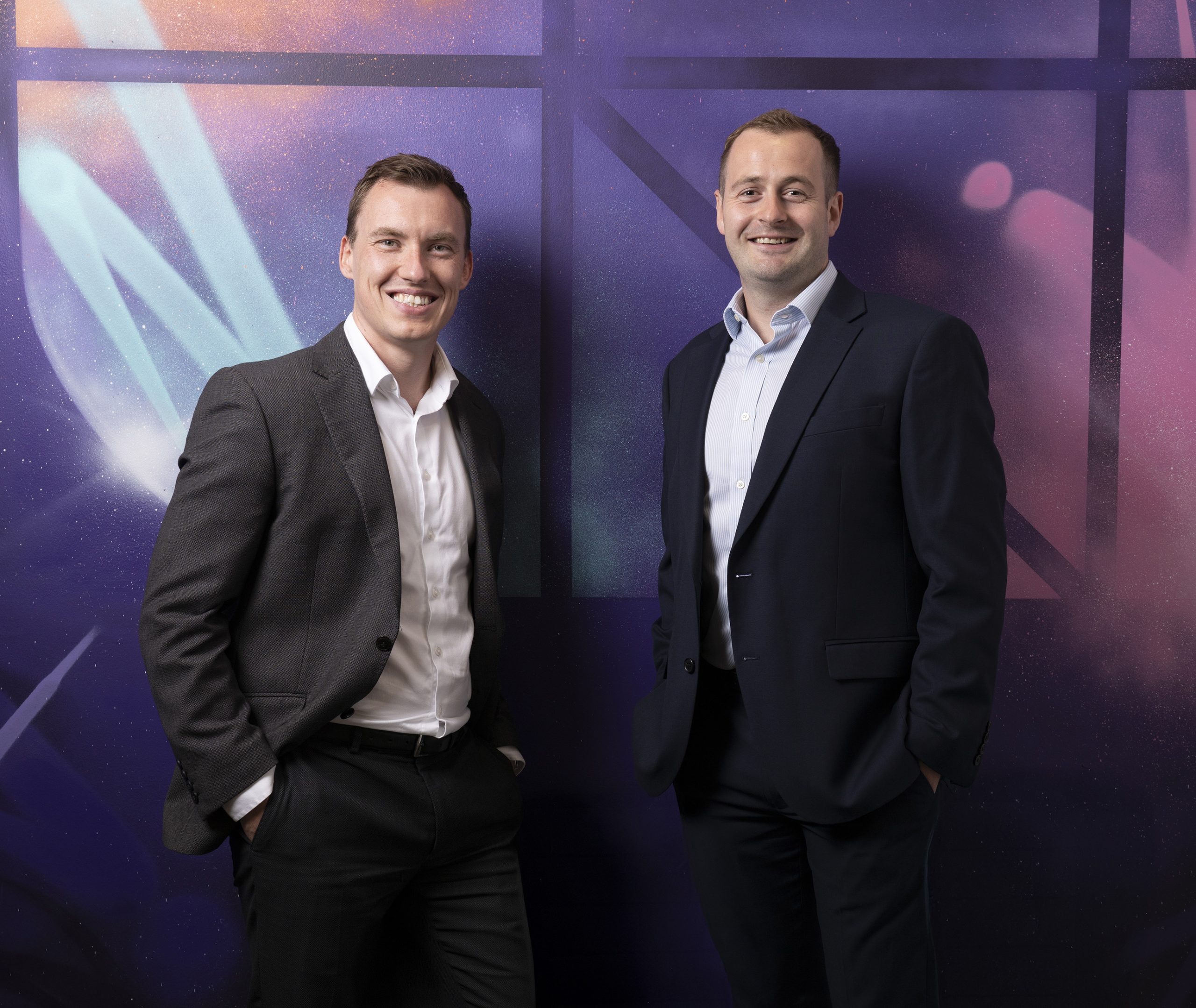 Danny and Stephen stood together against a colourful spray painted wall smiling to camera.