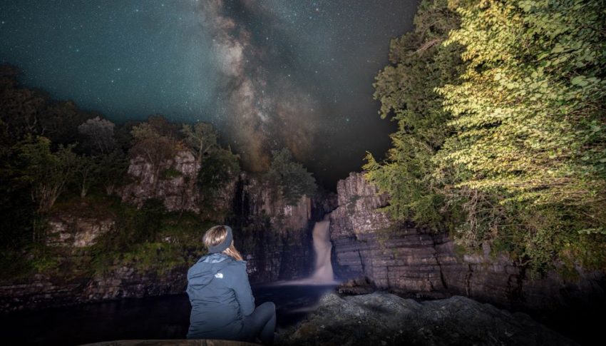 Person sat overlooking the High Force Waterfall with a starry sky overhead.