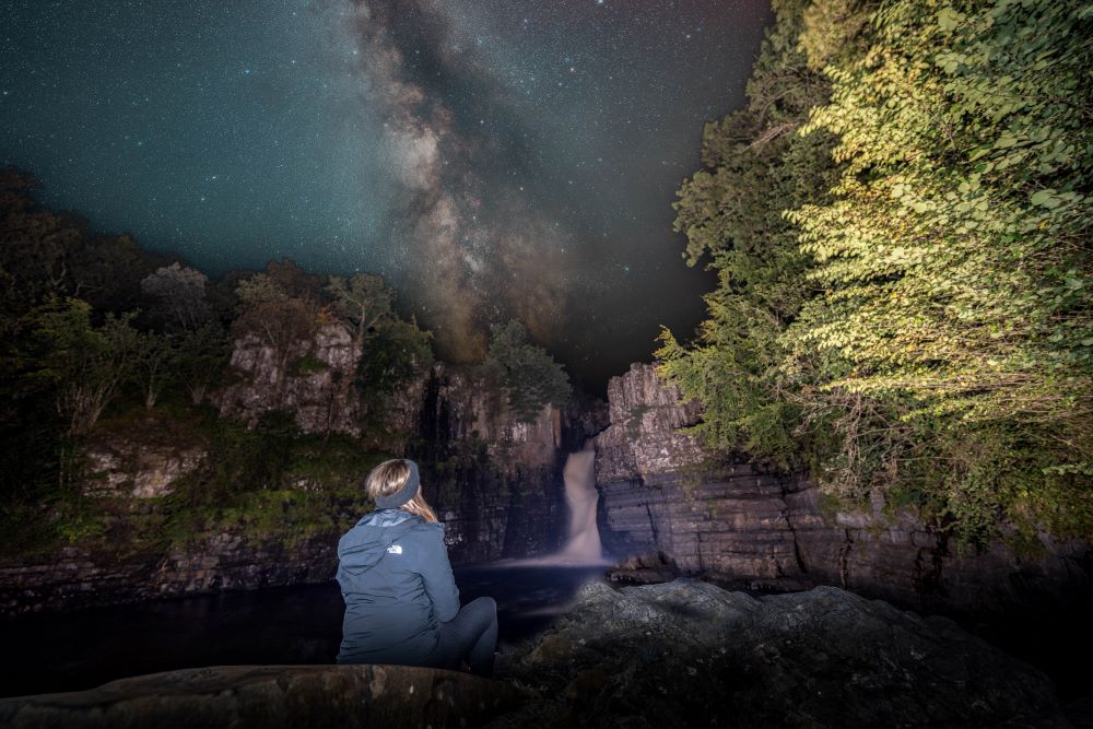 Person sat overlooking the High Force Waterfall with a starry sky overhead.