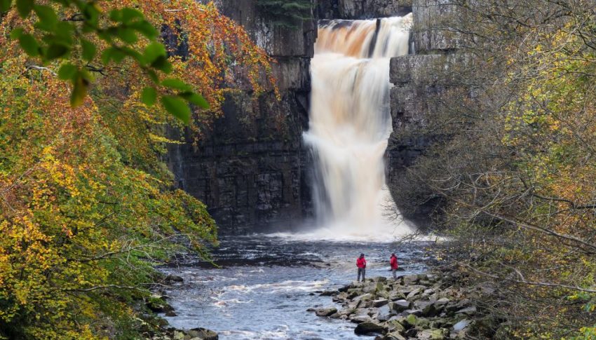 Large waterfall surrounded by trees with a rocky river bank where two people in red jackets are looking up at the waterfall.