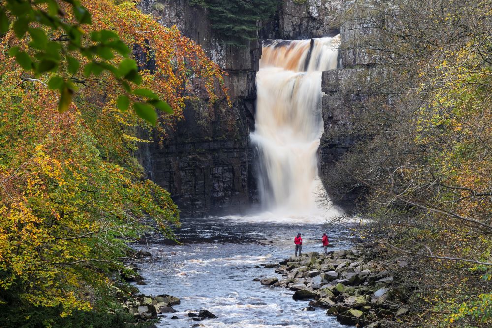 Large waterfall surrounded by trees with a rocky river bank where two people in red jackets are looking up at the waterfall.