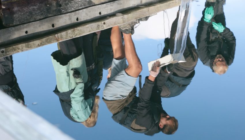 Reflection in water of a group of people and one is pouring water from a plastic container onto what is presumed to be the ground.