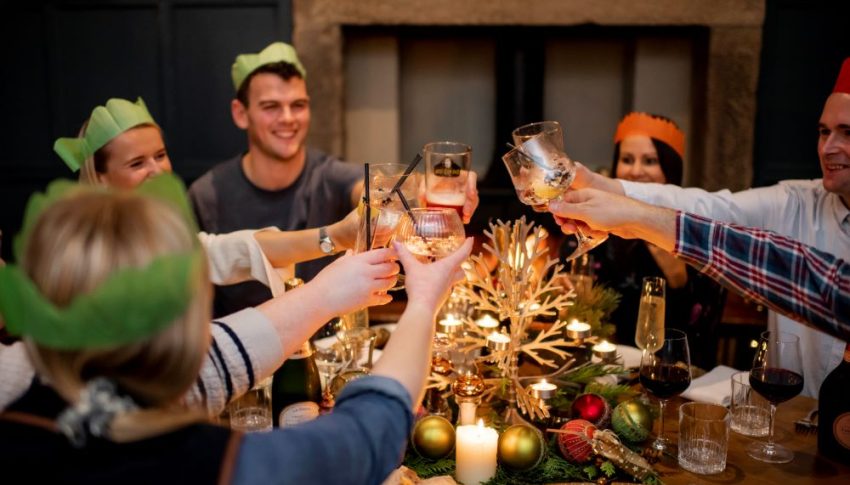 Group of people toasting their drinks around a festive-decorated table all wearing Christmas party hats.