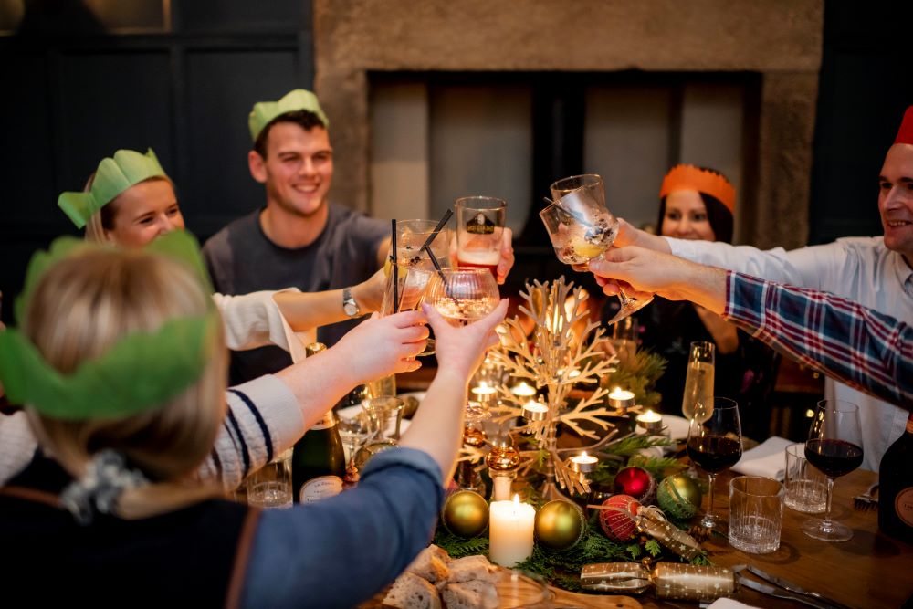 Group of people toasting their drinks around a festive-decorated table all wearing Christmas party hats.