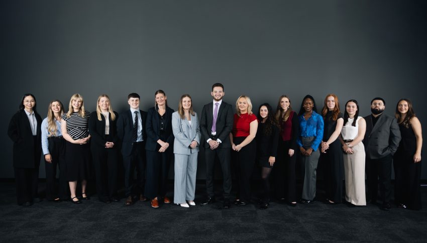 Group of 16 people stood in a line smiling to camera in a room with a black carpet and plain black wall.