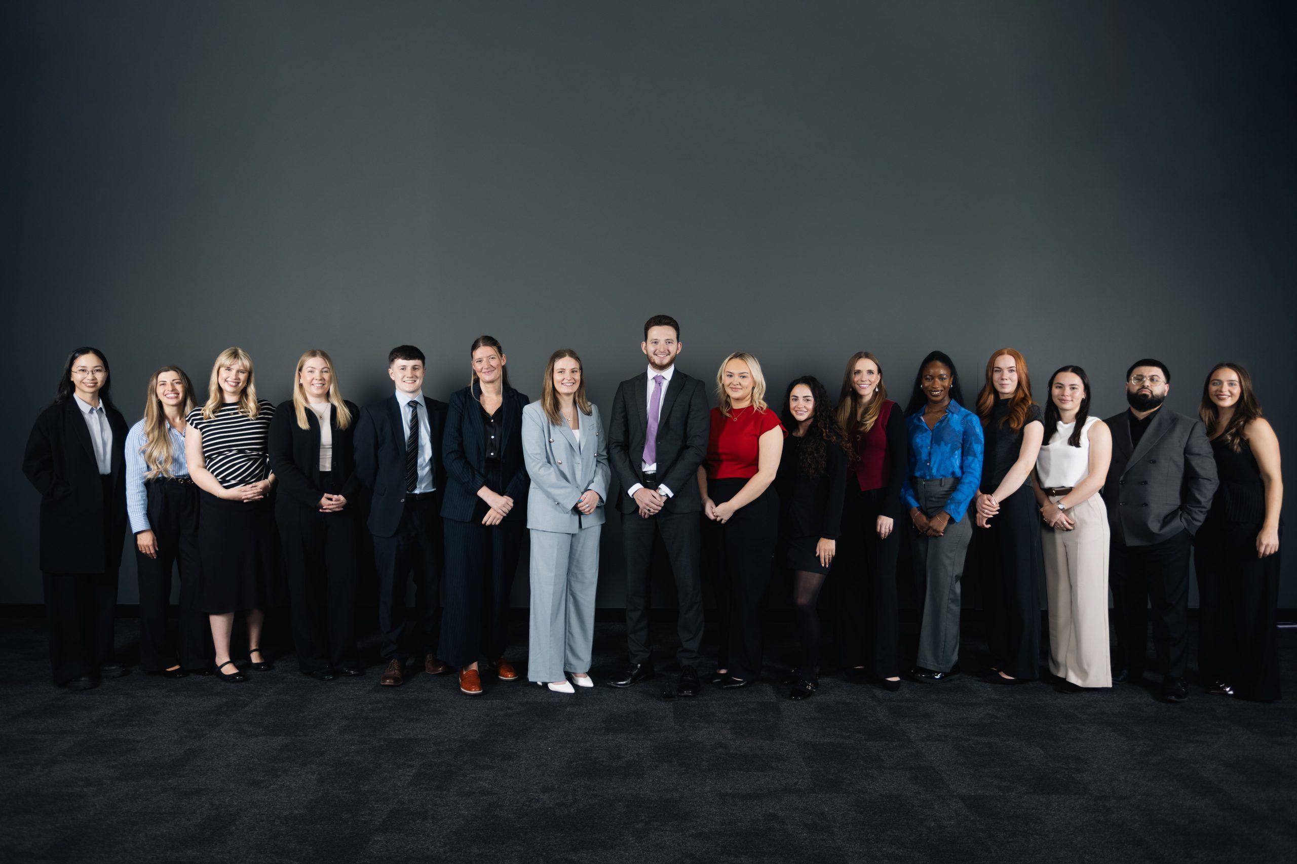 Group of 16 people stood in a line smiling to camera in a room with a black carpet and plain black wall.