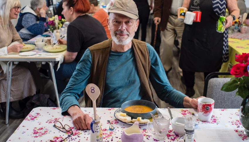 Person sat at a table enjoying a bowl of soup and smiling to camera.