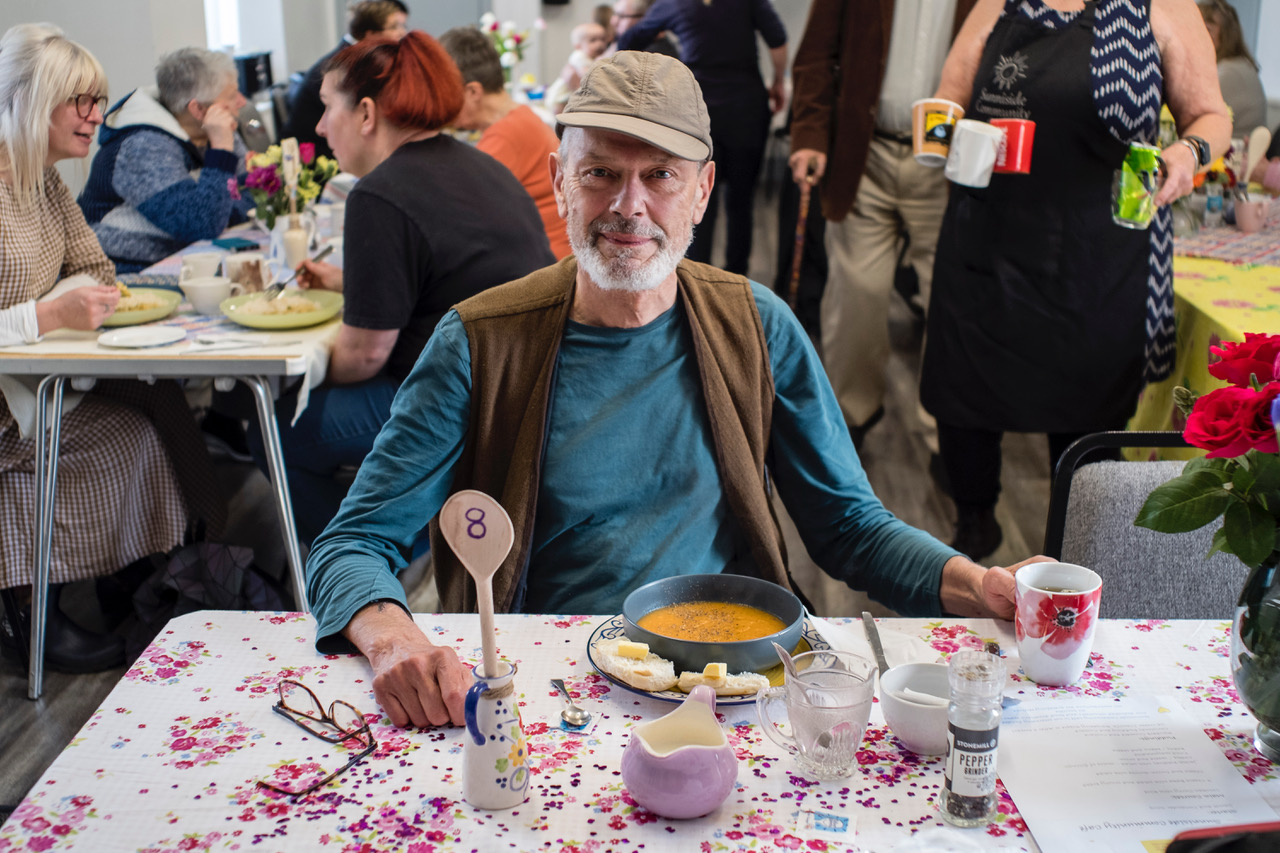 Person sat at a table enjoying a bowl of soup and smiling to camera.
